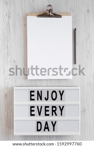 'Enjoy every day' words on a lightbox, clipboard with blank sheet of paper on a white wooden background, top view. Overhead, from above, flat lay. Copy space.