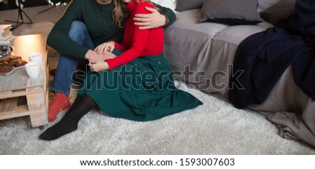 a happy, loving couple is sitting near the sofa, next to it is a Christmas tree, a table and tangerines, cups with chocolate, coffee with milk, cocoa, hugs. tenderness and comfort in the house