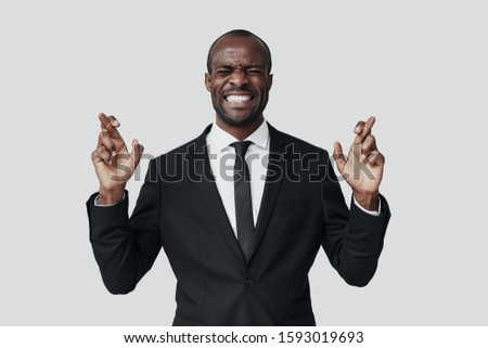 Handsome young African man in formalwear making a face and keeping fingers crossed while standing against grey background                