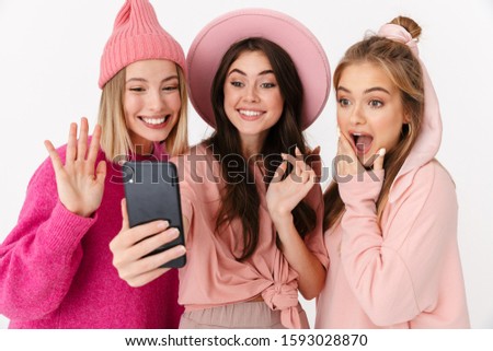 Image of three beautiful girls wearing pink clothes smiling and taking selfie photo on cellphones isolated over white background
