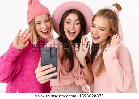 Image of three beautiful girls wearing pink clothes smiling and taking selfie photo on cellphones isolated over white background