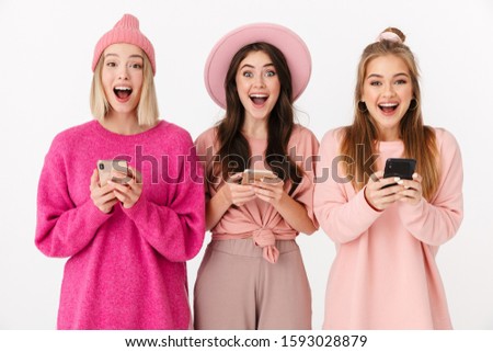 Image of three positive girls wearing pink clothes laughing and holding cellphones isolated over white background