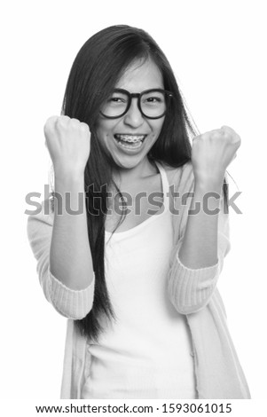 Studio shot of young happy Asian teenage girl smiling and looking motivated with both arms raised