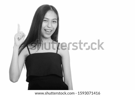 Studio shot of young happy Asian teenage girl smiling while pointing finger up
