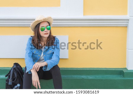 Beautiful smile Asian woman tourist in jean jacket and straw hat sitting on footpath with backpack on her side, looking something out next the yellow wall, travel and vacation concept.