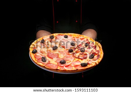 male cook in a black tunic and gloves holds a fresh pizza with olives in front of him
