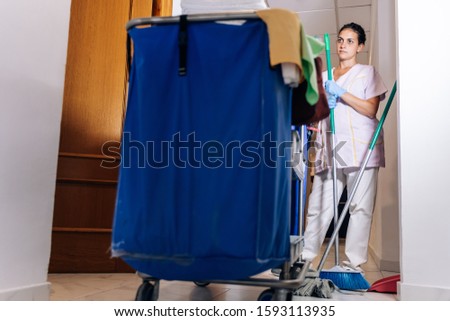 Stock photo of a Cleaning trolley and a hotel cleaner next door with a broom in hand. Lifestyle