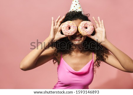 Image of amusing african american woman in party cone making fun with donuts isolated over pink background