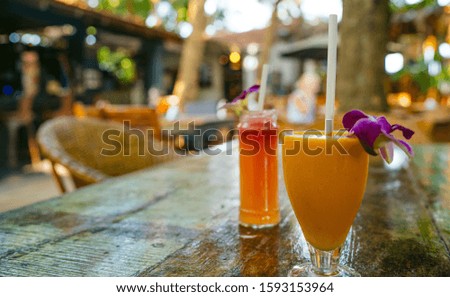 A healthy orange smoothie with a red smoothie in the background in a tropical cafe. The cool fresh juices or cocktails with straws and decorative flowers on the table.