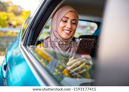 Muslim business woman in car. Writing a message on her mobile phone. Positive pensive Islamic woman in hijab sitting on backseat of taxi and drinking coffee while using gadget