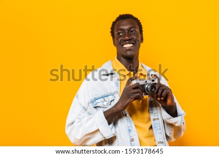Image of young african american guy in denim jacket smiling and photographing on retro camera isolated over yellow background