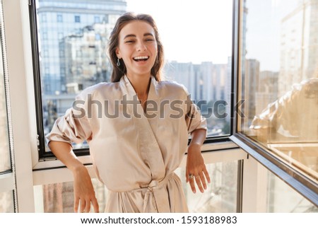 Image of laughing happy amazing young woman indoors posing at home in silk robe near big windows. Royalty-Free Stock Photo #1593188983