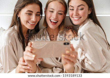 Image of a cheery pleased positive young girls women friends indoors on bed at the hen party at home take a selfie by mobile phone.