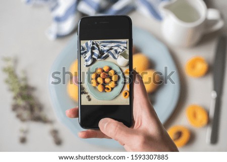 Female hand takes pictures on the phone Fresh organic apricots, whole and half with bone on a blue plate on a light gray background