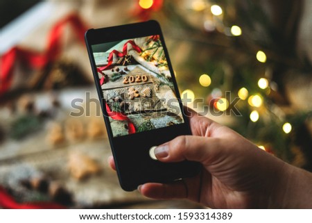 Female hand takes a photo on the phone of christmas cookies in the form of numbers 2020 on a wooden table with fir tree branches decoration and bokeh lights.