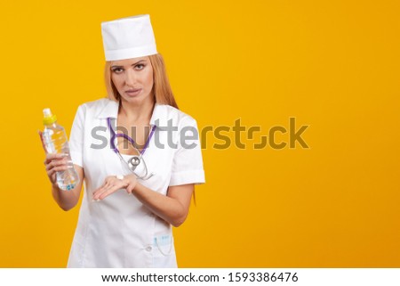 Emotional young woman doctor in a white coat and hat with a big white tablet and a bottle with drinking water in hands on a yellow background