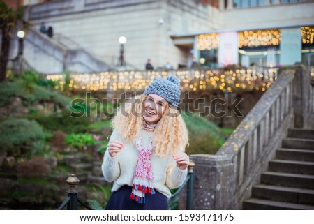 Portrait of smiling young curly blond woman in warm white natural sweater and blue knitted beanie hat with pompom looking at camera with the green park background and bokeh abstract lights