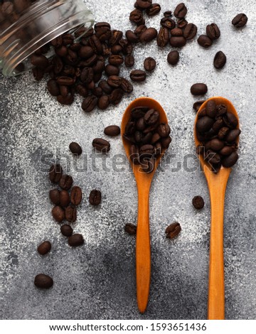 Coffee beans on two respective wooden spoon with a grey rough background , with a spilled jar of beans