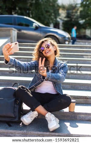 Portrait of pretty young woman wearing headphones taking selfie photo on smartphone while sitting on stairs outdoors