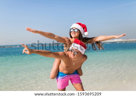 Christmas vacation beach couple doing piggyback fun. Paradise beach travel bikini woman having fun man carrying girl laughing in christmas santa hat.