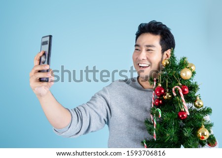 Asian man wearing gray sweater holding  christmas tree and taking photo on blue background.