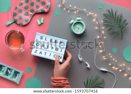Healthy night sleep creative concept. Sleeping mask, alarm clock, earphones, earplugs, tea and pills. Split two tone pink craft paper background with light garland. Text "sleep optimization in hand.