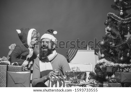 Santa and little assistant among gift boxes by Christmas tree. Christmas family opens presents on dark red background. Winter holiday concept. Man with beard and cheerful face laughs together with son