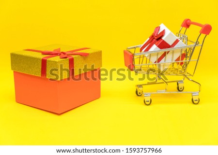 gift box in shopping cart and big gift box on yellow background next to red hearts, copy space. Gift concept, Valentine's Day, holiday