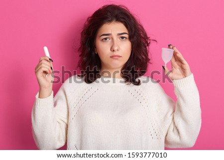 Picture of upset unsatisfied brunette having unpleasant facial expression, holding tampon and menstrual cup in both hands, standing isolated over pink background in studio. Critical days concept.