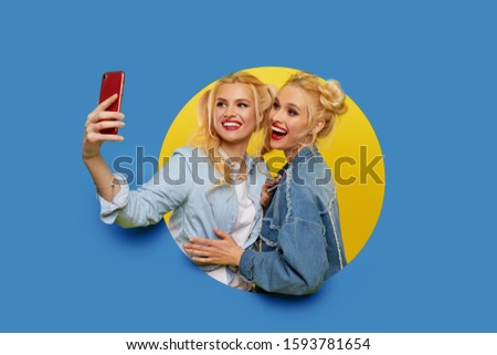 Image of two young happy girlfriends women standing over a blue background. Twins peep out of a hole in the wall. Looking to the side to take a selfie on the mobile phone.