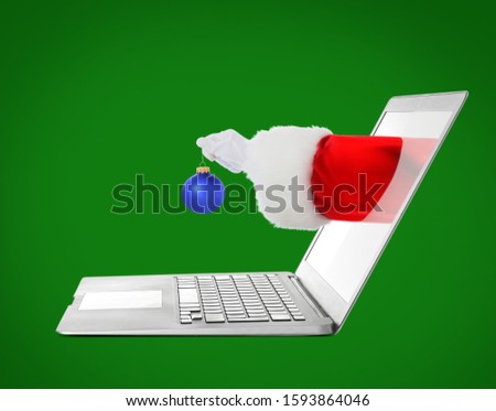 Santa sticking hand with Christmas ball out of laptop screen against color background