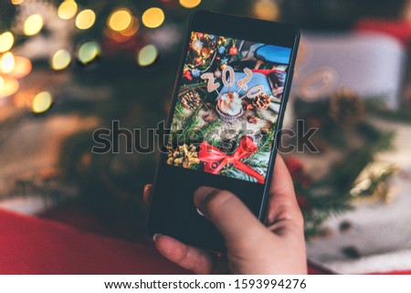Female takes a picture on the phone of christmas cupcake, fir tree branches decoration and bokeh lights. Close up.