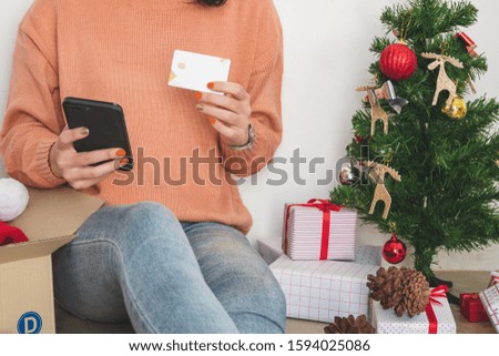 young woman in sweater sit on floor, using mobile phone, and holding credit card for christmas online shopping