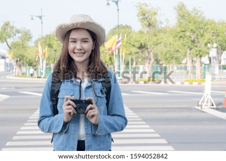 A young Asian traveler stands happily smiling on the street at the crosswalk, holding a camera, wearing a white T-shirt, covered with a denim jacket. It takes a closed up.