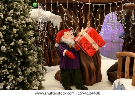 Under the beautifully decorated Christmas tree, a gnome sits and looks at a big red present. Christmas card with a gnome.