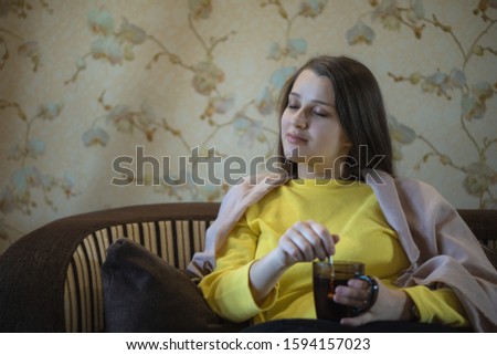 A picture of nice and thoughtful girl sits at sofa and drinks tea. She has brown blanket on her shoulders. Woman got cold. She suts at window