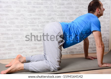 Full length portrait of attractive man working out at home in living room, doing yoga or pilates exercise on mat. Cow posture, asana paired with Cat Pose on the exhale. Side view