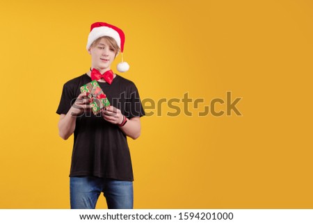Young guy in a red santa hat holds a gift box in his hands and poses on a yellow background