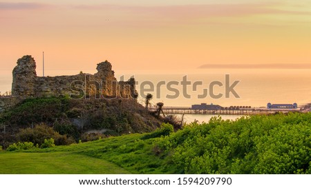 Historical Hastings castle set on a hill with the sea in the background. Bathed in the soft orange yellow glow of the sinking sun. Hastings Pier is also visible, as is Beachy Head in the far distance. Royalty-Free Stock Photo #1594209790