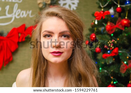 Portrait of a girl with bright red lips, blond long hair against the background of a New Year tree. Young girl in a white warm coat, shoulders are visible. Holidays. Merry Christmas