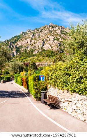Sideway of a road with wooden bench, dead end road sign, shaped hedge on stone fence and private houses, hotels of a beautiful city Limone Sul Garda with high dolomite mountains on the background