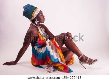 Studio fashion portrait of young African woman in summer dress and ethnic head wrap, white background, sitting girl, high heels