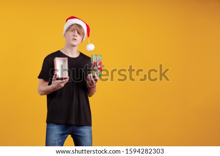 Young guy in a red santa hat holds a gift box in his hands and poses on a yellow background