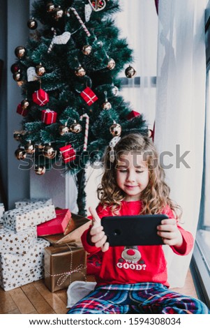 portrait of beautiful kid girl at home by the christmas tree and using mobile phone