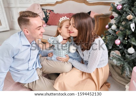 Loving family. Mom and dad hugging little daughter . Parents and baby child having fun near Christmas tree and white fireplace indoors. Merry Christmas and Happy New Year. Cheerful pretty people.