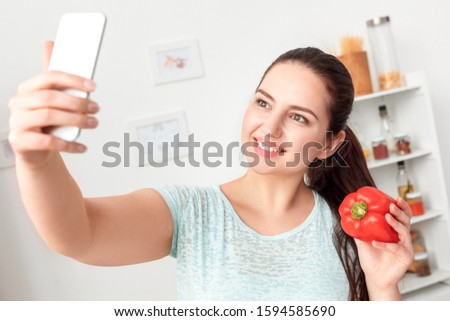 Young chubby woman standing in kitchen taking selfie photo on smartphone poing to camera holding bell pepper smiling happy close-up