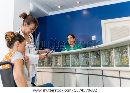 Stock photo of a girl with wheelchair talking to a doctor with a serious expression with a radiography in the hand in the reception of a hospital and a nurse in the background in the office