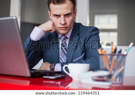 Young businessman working in bright office, sitting at desk with laptop. Expression of emotions.