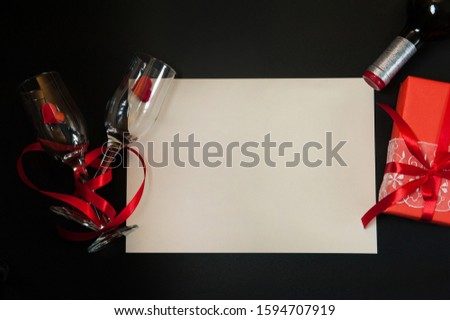 The Concept Of Valentine's Day. Paper blank, two narrow empty glasses intertwined with a red ribbon, a gift box, red hearts and a bottle of wine on a black background. Free space for your text.