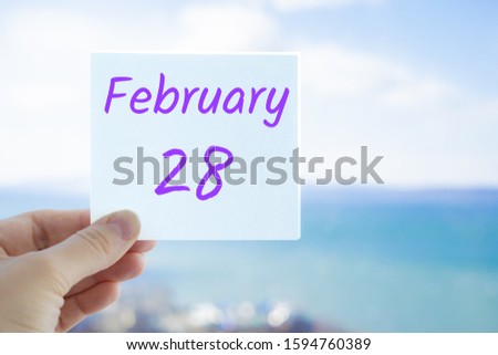 February 28th. Hand holding sticker with text February 28 on the blurred background of the sea and sky. Copy space for text. Month in calendar concept